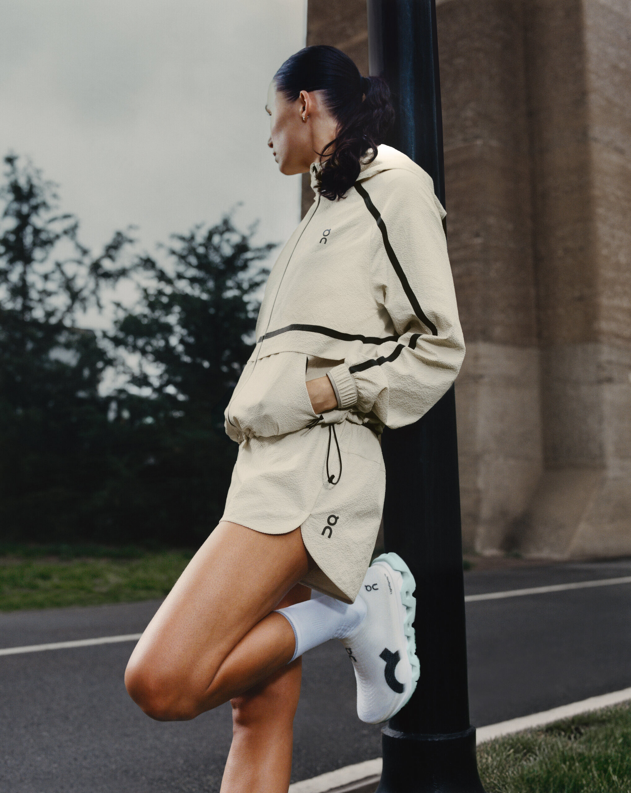 A runner in a cream On jacket and shorts stretching one leg against a lamppost beside a road